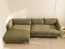 Load image into Gallery viewer, An olive green sectional sofa with a textured, dotted fabric pattern, positioned in a minimalist room with a white brick wall and light-colored rug.