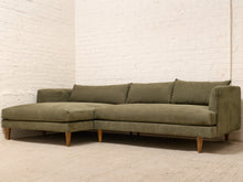 Load image into Gallery viewer, A mid-century modern sectional sofa in olive green fabric with a subtle geometric pattern and light wood legs, set against a white brick wall and light concrete floor.
