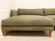 Load image into Gallery viewer, A mid-century modern sectional sofa upholstered in olive green and beige checkered fabric, featuring tapered wooden legs, set against a white brick wall and light-colored rug.