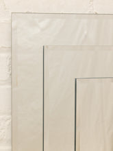 Load image into Gallery viewer, A close-up of a large rectangular mirror with a thin silver frame, leaning against a textured off-white brick wall. The composition is cropped to show the reflective surface and the edge of the frame.