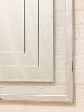 Load image into Gallery viewer, A close-up of a large rectangular mirror with a slim silver frame, reflecting a white paneled cabinet against a textured off-white brick wall.