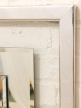 Load image into Gallery viewer, A close-up of a large rectangular mirror with a thick, polished silver frame, leaning against a textured white brick wall.