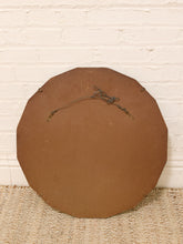 Load image into Gallery viewer, The back of a round, octagonal brown wall hanging or sign with a metal hanging wire and two small metal loops, leaning against a white painted brick wall on a woven jute rug.