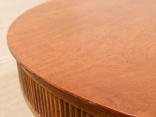 Load image into Gallery viewer, Close-up of a round wooden table top with a warm reddish-brown finish and visible wood grain, showing part of the fluted base.