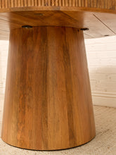 Load image into Gallery viewer, A close-up of a rustic wooden table base with a tapered, flared design and visible wood grain, set against a white brick wall and light-colored carpet.