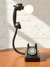 Load image into Gallery viewer, A vintage-style black rotary telephone turned into a lamp, with the handset detached and connected to a glowing white globe bulb, set on a wooden table against a white brick wall.