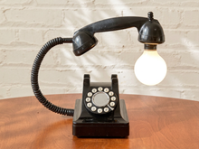 Load image into Gallery viewer, A novelty lamp shaped like a vintage black rotary telephone, with the handset turned into a light bulb, sitting on a wooden table against a white brick wall.