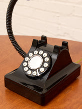 Load image into Gallery viewer, A glossy black vintage rotary telephone with a coiled cord, resting on a wooden tabletop against a light brick wall.