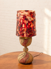Load image into Gallery viewer, A decorative table lamp with a marbled red, orange, and cream patterned shade and a matching sculptural base, displayed on a wooden table against a white brick wall.