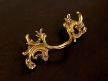 Load image into Gallery viewer, An ornate baroque-style gold metal drawer pull handle with intricate scrollwork, displayed on a dark brown wooden surface.