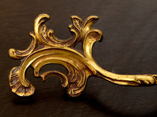 Load image into Gallery viewer, An ornate gold-toned metal decorative piece with intricate scrolling leaf and scrollwork details, photographed close-up on a dark brown wooden surface.