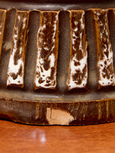 Load image into Gallery viewer, Close-up of a dark brown ceramic or stone object with vertical ridges and irregular white marbled patches, resting on a warm brown wooden surface.