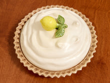 Load image into Gallery viewer, A ceramic pie dish filled with white cream or whipped icing, decorated with a small yellow lemon and green leaf on top, set on a wooden surface.