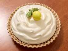 Load image into Gallery viewer, A lemon pie with a thick white custard-like filling and a fluted brown crust, topped with a small yellow lemon and green leaf decoration, set on a wooden surface.