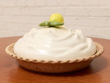 Load image into Gallery viewer, A ceramic lidded pie dish with a creamy white top and a small green leaf detail topped with a yellow sphere, set on a wooden table against a white brick wall.