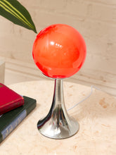 Load image into Gallery viewer, A modern table lamp with a glossy translucent orange-red spherical globe on a polished silver metal pedestal base, shown on a light stone tabletop with books and a leaf in the background.