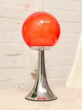 Load image into Gallery viewer, A modern table lamp with a glossy orange-red spherical shade on a polished chrome flared base, set on a light stone surface against a white brick wall.