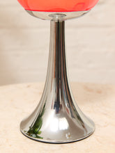 Load image into Gallery viewer, A close-up of a modern pedestal chalice or goblet with a reflective silver metal stem and base, holding a translucent coral-red bowl, set on a light beige surface.