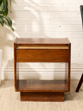 Load image into Gallery viewer, A mid-century modern wooden nightstand with a warm medium-brown finish, featuring a single drawer with a pull-out top shelf, set against a white brick wall and beige woven rug.