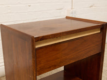 Load image into Gallery viewer, A mid-century modern wooden nightstand with a warm brown finish, featuring a single drawer with a light wood pull and an open lower shelf, set against a white brick wall background.