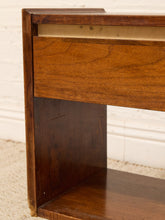 Load image into Gallery viewer, Close-up of a wooden furniture piece, showing a medium brown drawer front, dark brown side panel, and a light natural wood inner shelf against a white brick wall and beige carpet.