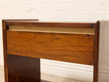 Load image into Gallery viewer, Close-up of a wooden furniture piece, showing a medium-to-dark brown wood grain drawer front with a light beige inner panel and a dark brown frame, set against a white brick wall background.