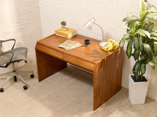 Load image into Gallery viewer, A mid-century modern wooden desk with a warm brown finish, styled in a room corner with a white brick wall, beige carpet, a yellow rotary phone, a silver desk lamp, and a potted plant.