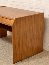 Load image into Gallery viewer, A close-up of a mid-century modern wooden desk with a warm brown teak wood finish, featuring a drawer and open shelf, set against a white brick wall and beige carpet.