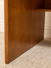Load image into Gallery viewer, Close-up of a mid-century modern wooden desk leg with a warm brown finish, standing on a textured beige carpet against a white brick wall.