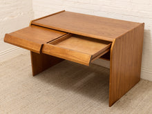 Load image into Gallery viewer, A mid-century modern wooden desk with a warm honey finish, featuring a top desk surface, a lower drawer with its front panel pulled out, and a side drawer partially extended. The desk is photographed in a room setting on a textured beige carpet against a white painted brick wall.