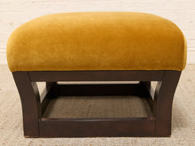 Load image into Gallery viewer, A square upholstered ottoman with a mustard yellow velvet cushion and a dark brown wooden frame, set against a white brick wall and light beige carpet.
