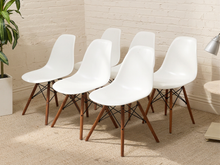 Load image into Gallery viewer, Six modern white molded plastic dining chairs with brown wooden legs, arranged in a semi-circle on a beige carpeted floor against a white brick wall.