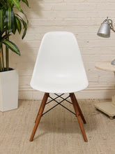 Load image into Gallery viewer, A modern white molded plastic side chair with wooden legs, positioned in a styled room setting with a brick wall, potted plant, and side table.
