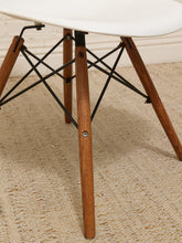 Load image into Gallery viewer, Close-up of a modern chair leg with a white molded seat, medium brown wooden legs, and a black metal wire support frame on a textured beige rug.