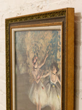 Load image into Gallery viewer, A framed impressionist painting of two ballerinas, shown hanging on a white brick wall. The artwork features soft pastel tones of blue, green, and cream, enclosed in an ornate gold-toned frame with a white mat.