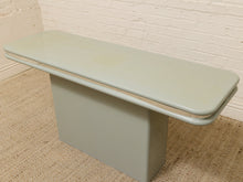 Load image into Gallery viewer, A mid-century modern console table with a light sage green painted metal frame and a matching rectangular seat, set on a woven jute rug against a white brick wall.