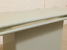 Load image into Gallery viewer, A close-up of a modern console table featuring a glossy pale sage green painted base and a clear glass top with a thin metallic trim.