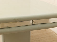 Load image into Gallery viewer, Close-up of a light beige or off-white table with a polished metallic silver apron and edge trim, showing some wear and scuffs on the surface.