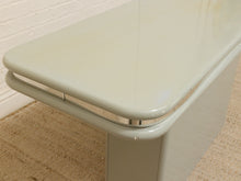 Load image into Gallery viewer, Close-up of a light sage green or off-white table with rounded corners and a glossy finish, showing part of the tabletop and leg on a beige textured rug against a white brick wall.