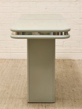 Load image into Gallery viewer, A mid-century modern side table with a light sage green painted metal base and a matching rounded rectangular top, accented by a polished chrome band, set against a white brick wall and beige rug.