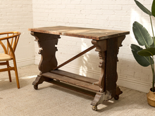 Load image into Gallery viewer, An antique dark wood writing desk with ornate turned legs and caster wheels, set against a white brick wall with a potted plant and a wooden chair partially visible.