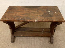 Load image into Gallery viewer, A rustic dark brown wooden table with a distressed finish, featuring a rectangular top, decorative turned legs, and a lower support rail, set on a textured beige rug against a white brick wall.