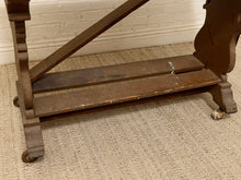 Load image into Gallery viewer, Close-up of the lower section of a dark brown wooden table or stand with casters, resting on a textured beige rug against a white brick wall.