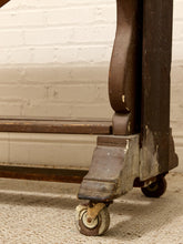 Load image into Gallery viewer, Close-up of the lower corner of a dark brown wooden rolling table or cart with casters, showing the leg joint, worn wood finish, and caster wheel on a carpeted floor against a white brick wall.
