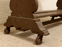 Load image into Gallery viewer, Close-up of a dark brown wooden wheeled bench or stool with a distressed finish, resting on a textured beige rug against a white brick wall.