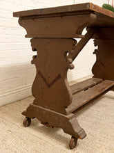 Load image into Gallery viewer, A rustic dark brown wooden console table with ornate carved legs and casters, shown in a room setting on a textured rug against a white brick wall.