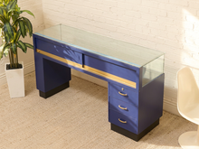 Load image into Gallery viewer, A modern navy blue vanity desk with a clear glass top, gold trim, and three drawers on the right side, set against a white brick wall and beige carpet.
