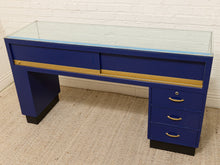 Load image into Gallery viewer, A blue wooden writing desk with a glass top, gold drawer pulls and trim, and black feet, photographed in a room with a white brick wall and beige carpet.