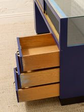 Load image into Gallery viewer, Close-up of a mid-century modern console table with a deep blue painted frame, a glass top, and three open wooden drawers pulled out, set on a beige carpeted floor.