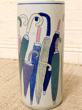 Load image into Gallery viewer, A tall cylindrical ceramic vase or planter with a white base, decorated with a colorful pattern of stylized umbrellas in blue, green, purple, and cream. The vase is photographed standing on a beige woven mat against a white painted brick wall.
